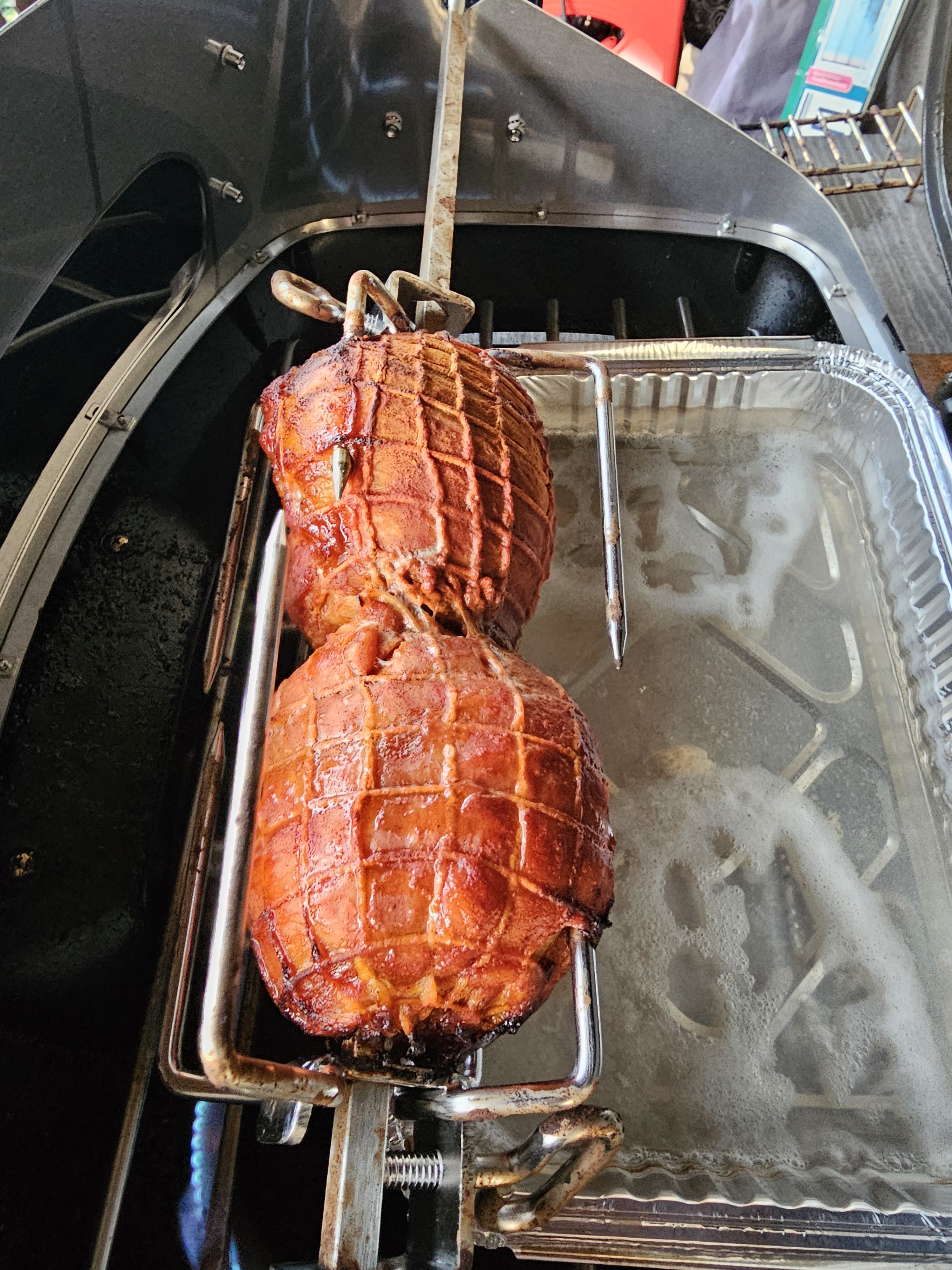 Zwei goldbraun gegrillte Fleischstücke in einem Netz auf einem Drehspieß über einer mit Wasser gefüllten Aluminiumschale in einem Gasgrill.