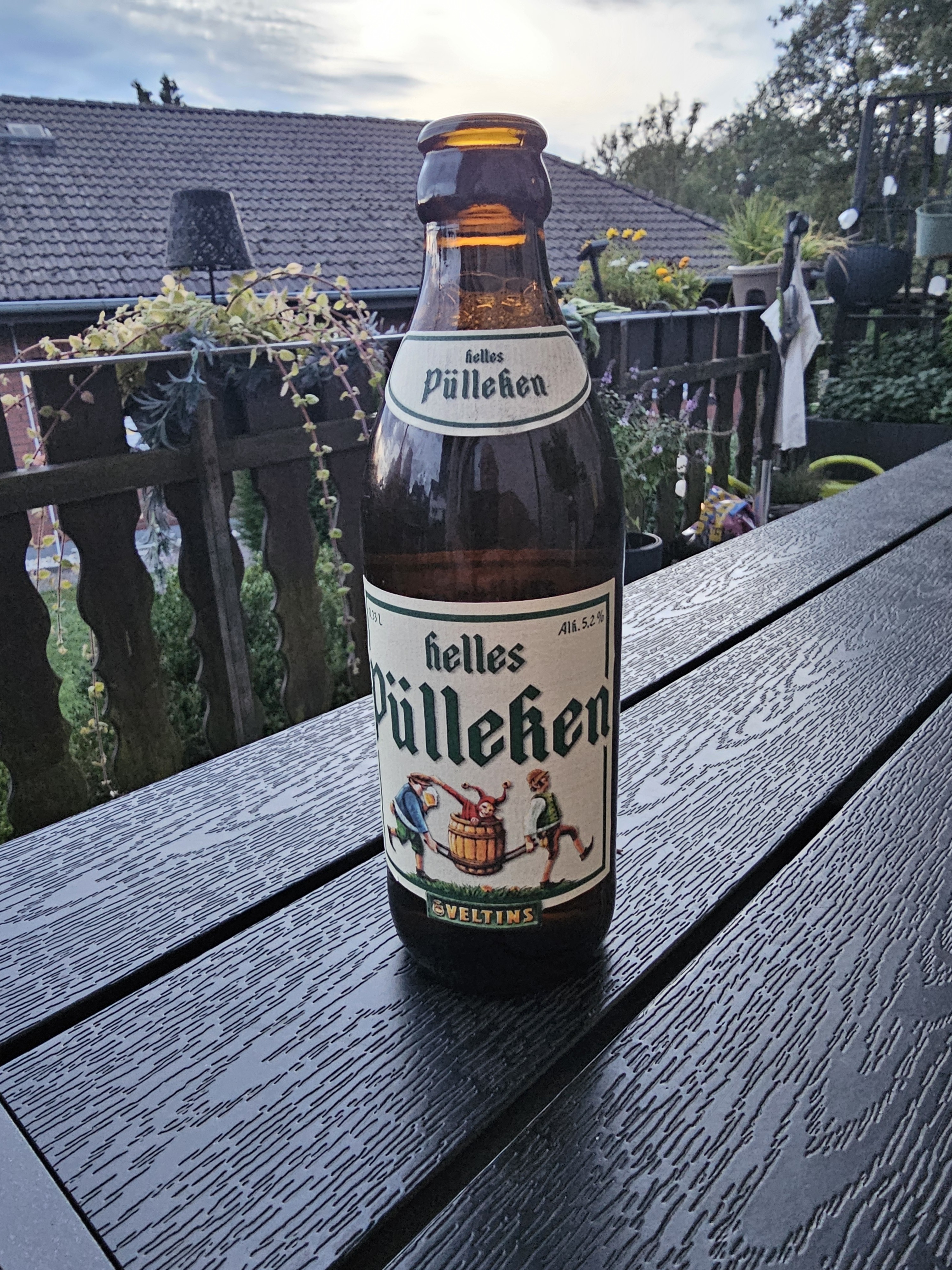 Bierflasche mit dem Etikett 'helles Pülleken' auf einem Holztisch vor einem Balkongeländer. Im Hintergrund dämmert der Abendhimmel