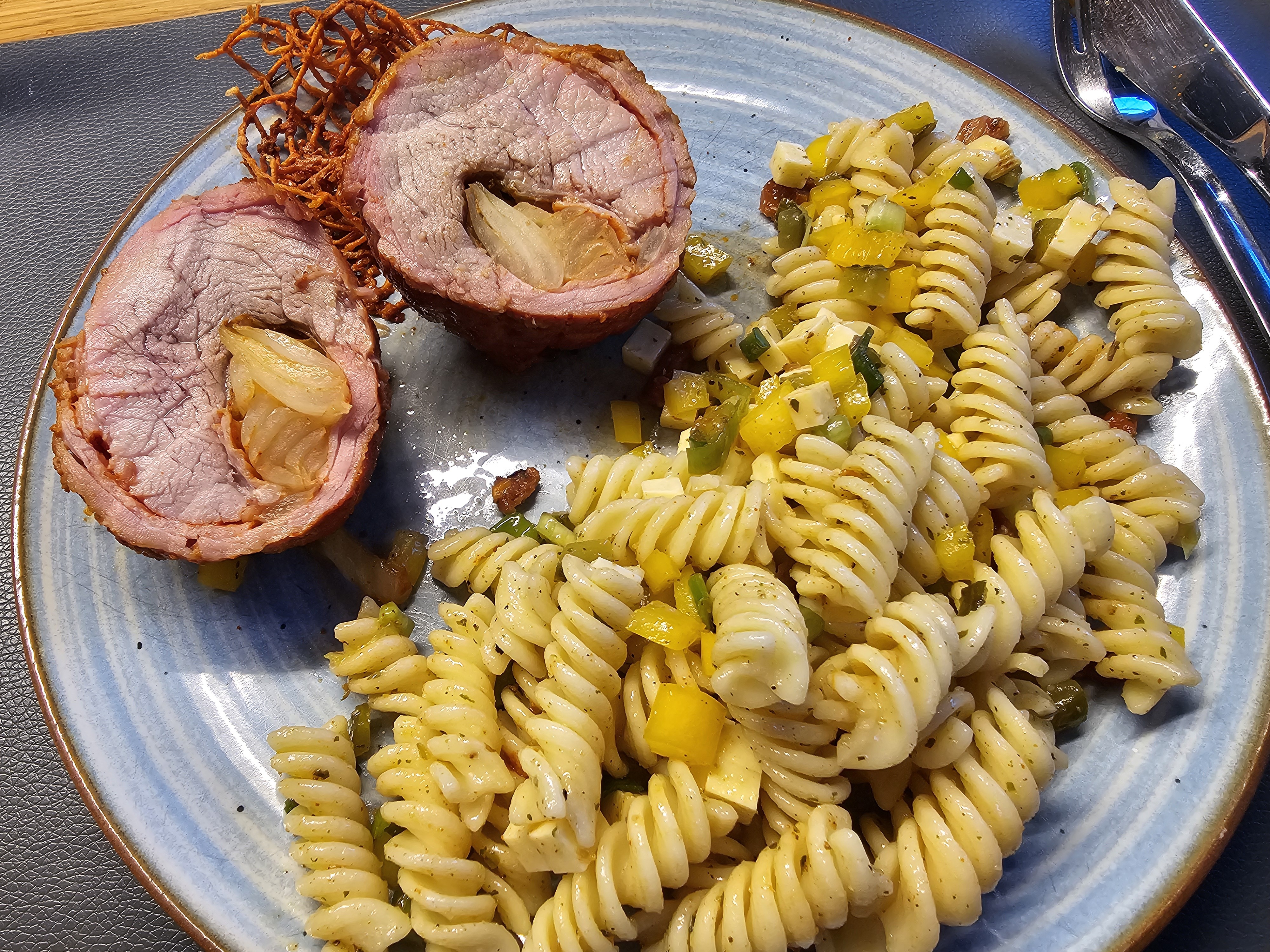 Zwei Scheiben gefüllter Schweinebraten mit Zwiebelkern neben einem Nudelsalat aus Fusilli, gelben Paprikastücken und Käsewürfeln auf einem blauen Teller.